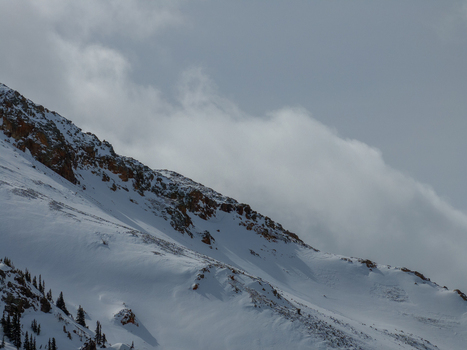 View of snow covered mountain peaks with rocky ridges and evergreen trees under a cloudy sky in Colorado United States.