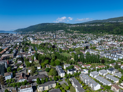 Aerial view of the urban landscape with residential buildings lush green parks and rolling hills under a clear blue sky in Biel Bienne Canton of Bern Switzerland.