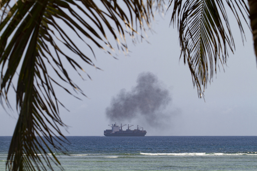 View of a large cargo ship emitting dark smoke on the horizon framed by tropical palm fronds at Tiwi Beach Tiwi Wilaya ya Kwale Kenya. View of a large cargo ship emitting dark smoke on the horizon framed by tropical palm fronds at Tiwi Beach Tiwi Wilaya ya Kwale Kenya.