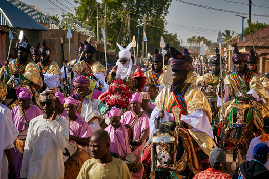Zaria city Nigeria 22 March 2026 View of a vibrant ceremonial procession of horsemen in resplendent golden attire and a crowd in vivid pink moving through bright streets. Zaria city Nigeria 22 March 2026 View of a vibrant ceremonial procession of horsemen in resplendent golden attire and a crowd in vivid pink moving through bright streets.