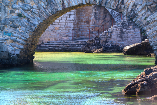 View of a historic stone archway over clear turquoise water with stone steps and rocky shores in Cascais Lisbon Portugal.