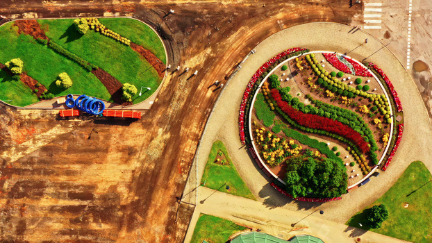 Aerial view of a circular roundabout featuring vibrant flower beds with red yellow and green plants surrounded by gravel paths and patches of grass in Latvija. Aerial view of a circular roundabout featuring vibrant flower beds with red yellow and green plants surrounded by gravel paths and patches of grass in Latvija.