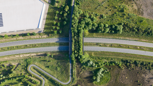 Aerial view of a green wildlife crossing bridge over a highway surrounded by lush vegetation grassy fields and a large industrial facility with solar panels in Venlo Limburg Netherlands. Aerial view of a green wildlife crossing bridge over a highway surrounded by lush vegetation grassy fields and a large industrial facility with solar panels in Venlo Limburg Netherlands.