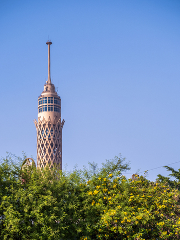 View of a tall slender tower rising beyond the green and yellow foliage against a clear blue sky showcasing architectural grandeur Zamalek Cairo Governorate Egypt. View of a tall slender tower rising beyond the green and yellow foliage against a clear blue sky showcasing architectural grandeur Zamalek Cairo Governorate Egypt.