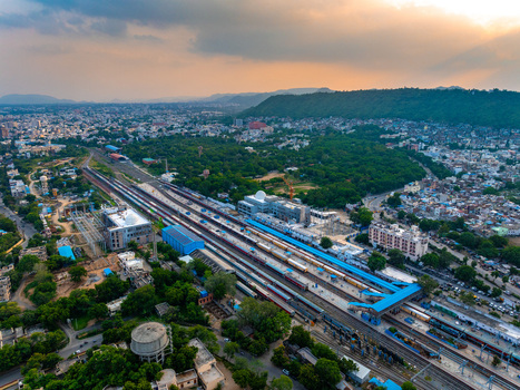 Aerial view of Udaipur railway station with trains at platforms surrounded by urban buildings trees and distant hills under a dramatic sunset sky in Udaipur Rajasthan India. Aerial view of Udaipur railway station with trains at platforms surrounded by urban buildings trees and distant hills under a dramatic sunset sky in Udaipur Rajasthan India.