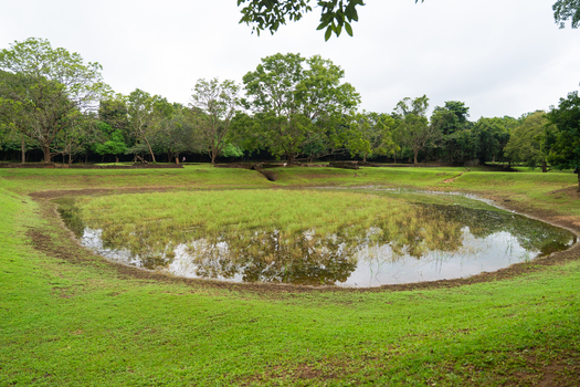 View of the ancient water gardens with a grassy pond surrounded by lush green trees and stone ruins in Sigiriya Central Province Sri Lanka. View of the ancient water gardens with a grassy pond surrounded by lush green trees and stone ruins in Sigiriya Central Province Sri Lanka.