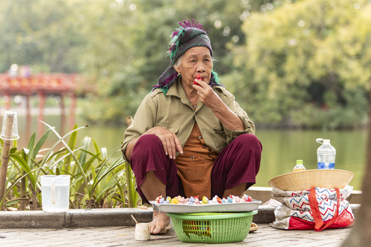 Hanoi Ha Noi Vietnam 11 November 2025 View of an elderly woman sitting by Hoan Kiem Lake selling colorful traditional dough figurines with a conical hat and water bottles nearby.