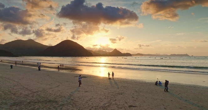 Aerial view of a sandy beach at sunset with people walking along the shore calm sea and distant mountains under a golden sky in Lombok West Nusa Tenggara Indonesia.