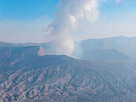 Aerial view of Mount Bromo with volcanic smoke rising from the crater rugged volcanic landscape ash covered slopes and clear blue sky in Tosari East Java Indonesia.