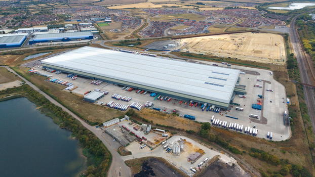 Aerial view of a massive logistics warehouse with numerous trucks and trailers parked at loading bays situated near a lake and railway tracks in Bedford England United Kingdom.