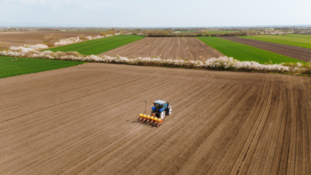 Aerial view of a blue tractor sowing seeds in the vast ploughed fields with a backdrop of green fields and white blossoming trees Sremska Mitrovica Vojvodina Serbia. Aerial view of a blue tractor sowing seeds in the vast ploughed fields with a backdrop of green fields and white blossoming trees Sremska Mitrovica Vojvodina Serbia.