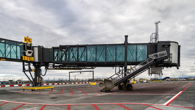 View of a modern glass walled jet bridge at an airport apron with red markings on the tarmac under a cloudy sky in Iceland.
