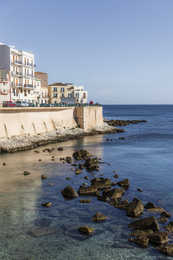 View of the coastline with white buildings perched above a stone wall overlooking the clear blue sea in Ortigia Sicily Italy.