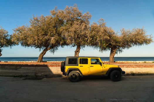 View of a yellow off road vehicle parked on a coastal road beside leafy trees and a low stone wall overlooking the Mediterranean Sea in Marsaskala Southern Region Malta. View of a yellow off road vehicle parked on a coastal road beside leafy trees and a low stone wall overlooking the Mediterranean Sea in Marsaskala Southern Region Malta.