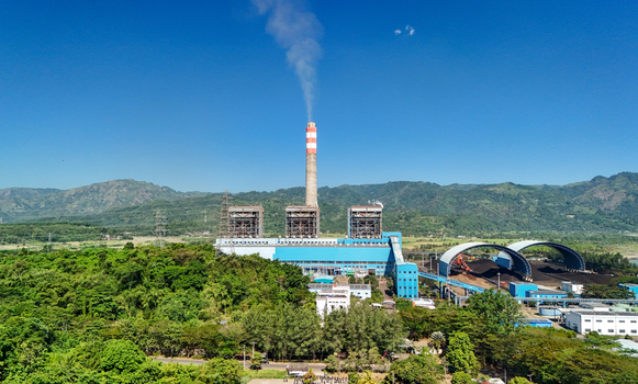 Aerial view of a power plant with a tall smokestack emitting smoke surrounded by lush green hills and trees under a clear blue sky in Indonesia.