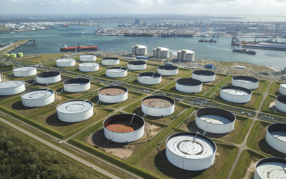 Aerial view of numerous white circular oil storage tanks in the Port of Rotterdam Netherlands arranged in a grid near a busy industrial harbor with cargo ships and cranes under a bright sky.