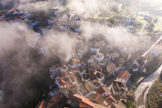 Aerial view of the historic village of Avo with red tiled rooftops partially obscured by thick morning mist and sunlight in Oliveira do Hospital Coimbra District Portugal.