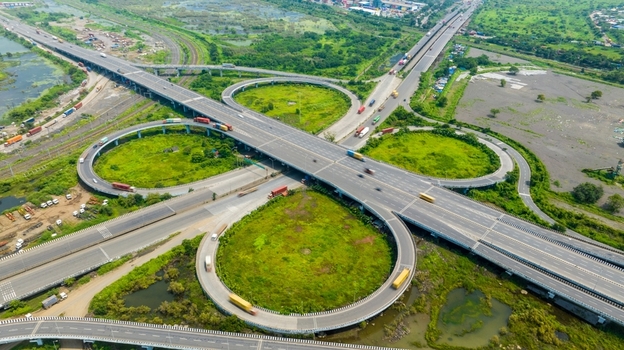 Aerial view of the JNPT Interchange featuring complex highway ramps and heavy truck traffic surrounded by lush green wetlands in Navi Mumbai Maharashtra India.