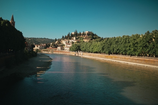 View of the Adige River flowing past tree lined banks with Castel San Pietro on the hill under a clear blue sky in Verona Veneto Italy.
