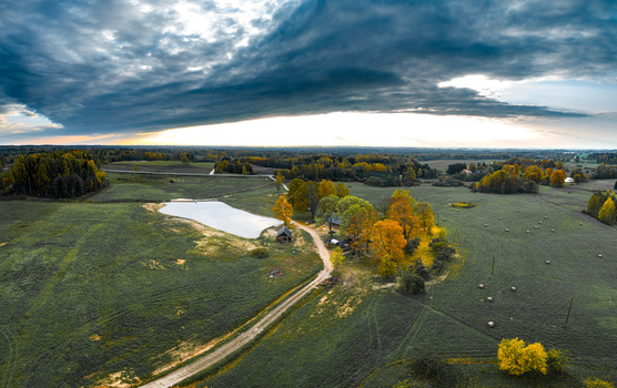 Aerial view of a rural landscape featuring a small pond a winding dirt road and vibrant autumn trees under a dramatic cloudy sky in Cesis Cesis Municipality Latvia. Aerial view of a rural landscape featuring a small pond a winding dirt road and vibrant autumn trees under a dramatic cloudy sky in Cesis Cesis Municipality Latvia.