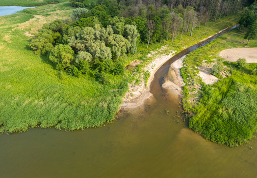 Aerial view of a small river flowing into a larger body of water surrounded by lush green vegetation trees and sandy banks in Tolkmicko Warmian Masurian Voiv Poland. Aerial view of a small river flowing into a larger body of water surrounded by lush green vegetation trees and sandy banks in Tolkmicko Warmian Masurian Voiv Poland.