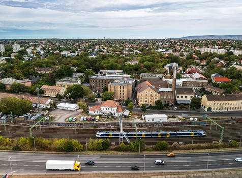 Aerial view of a passenger train on railway tracks beside a busy multilane road industrial buildings and leafy residential neighborhoods in Budapest Hungary in summer.