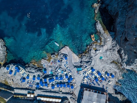 Aerial view of a rocky beach club with people relaxing under blue umbrellas and sunbeds along the turquoise coastline in Capri Campania Italy.