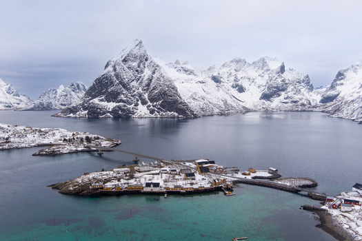 Aerial view of Hamnoy village in the Lofoten archipelago with yellow and red wooden houses on islands connected by bridges surrounded by snow capped mountains in Nordland Norway.