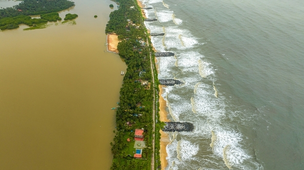 Aerial view of Mattu Beach a narrow strip of land separating calm backwaters from the wavy Arabian Sea with palm trees coastal houses and stone breakwaters in Udupi Karnataka India.