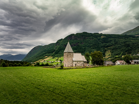 Aerial view of Hove Church surrounded by lush green fields and dramatic mountains under a cloudy sky in Vik i Sogn Vestland Norway.