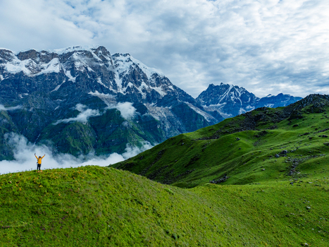 Aerial view of a hiker in a yellow jacket standing with arms raised on a lush green mountain ridge overlooking the snow capped Himalayan peaks and clouds in Nepal.