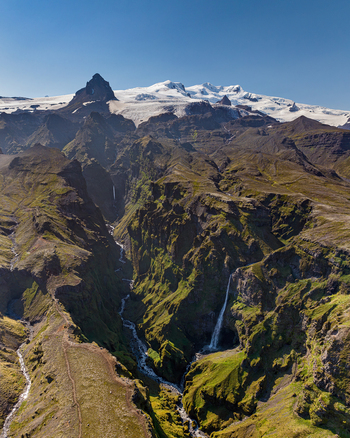 Aerial view of the deep Mulagljufur Canyon with its lush green mossy cliffs cascading waterfalls and snow capped mountains under a clear blue sky in Iceland. Aerial view of the deep Mulagljufur Canyon with its lush green mossy cliffs cascading waterfalls and snow capped mountains under a clear blue sky in Iceland.