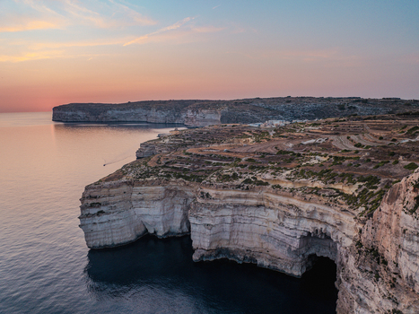 Aerial view of rugged cliffs meet the tranquil sea under a soft pastel sky where warm light kisses the horizon Munxar Gozo Malta. Aerial view of rugged cliffs meet the tranquil sea under a soft pastel sky where warm light kisses the horizon Munxar Gozo Malta.