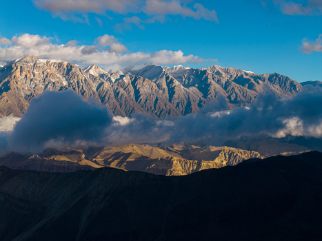 Aerial view of snow capped Himalayan mountain peaks with dramatic clouds and golden hour light illuminating the rugged slopes in Nepal.