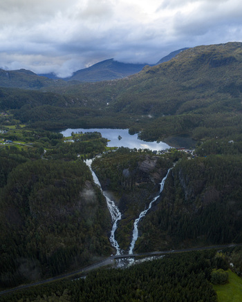 Aerial view of Latefossen waterfall cascading through dense green forest towards a valley road with a calm lake and misty mountains in the background Skare Vestland Norway.