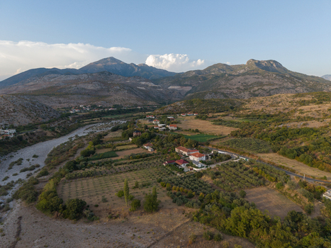 Aerial view of the rural landscape featuring a dry riverbed scattered houses olive groves and rugged mountains under a clear sky in Boks Qarku i Shkodres Albania.