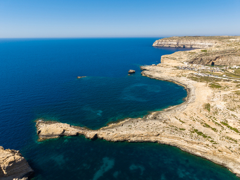 Aerial view of Dwejra Bay featuring rugged limestone cliffs and the deep blue Mediterranean Sea under a clear sky in Gozo Region Malta. Aerial view of Dwejra Bay featuring rugged limestone cliffs and the deep blue Mediterranean Sea under a clear sky in Gozo Region Malta.