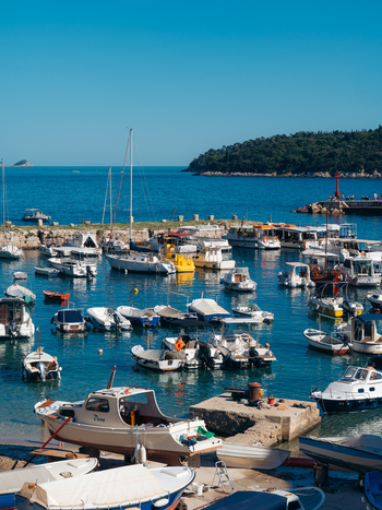 View of small boats moored in the harbor with people on the pier and lush green hills under a clear blue sky in Dubrovnik Dubrovnik Neretva County Croatia. View of small boats moored in the harbor with people on the pier and lush green hills under a clear blue sky in Dubrovnik Dubrovnik Neretva County Croatia.