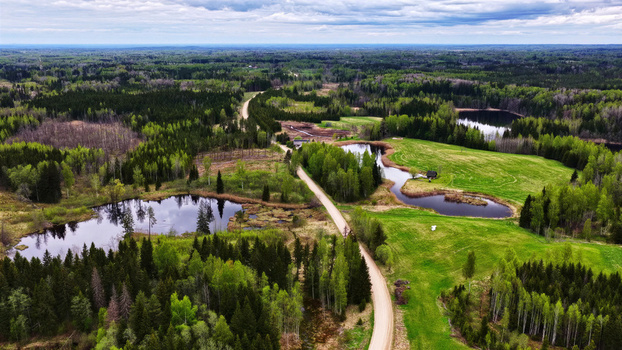 Aerial view of a winding dirt road cutting through lush green fields dense forests and small reflective ponds in the rural countryside of Latvia. Aerial view of a winding dirt road cutting through lush green fields dense forests and small reflective ponds in the rural countryside of Latvia.