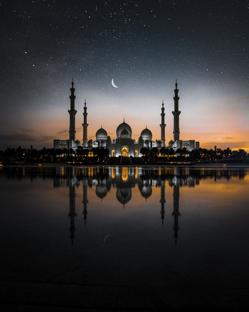 View of al zayed mosque illuminated at night with reflection in water and crescent moon Abu Dhabi United Arab Emirates.