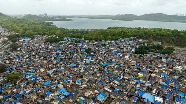 Aerial view of the dense Dharavi Slums featuring a vast collection of small rooftops narrow alleyways and proximity to a large lake and green hills in Mumbai Maharashtra India. Aerial view of the dense Dharavi Slums featuring a vast collection of small rooftops narrow alleyways and proximity to a large lake and green hills in Mumbai Maharashtra India.