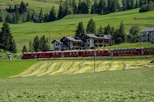 View of a red regional train passing through lush green meadows and alpine forests near the Sankt Gotthard Pass in Airolo Ticino Switzerland.
