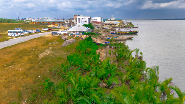 Aerial view of a waterfront area with traditional wooden boats moored along the shore lush palm trees and nearby buildings under a cloudy sky in Rivers Nigeria.