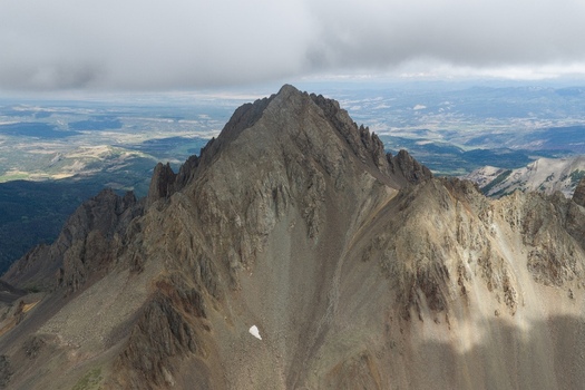 Aerial view of Mount Sneffels in the San Juan Mountains featuring rugged rocky slopes steep ridges and patches of snow under a cloudy sky in Ouray County Colorado United States. Aerial view of Mount Sneffels in the San Juan Mountains featuring rugged rocky slopes steep ridges and patches of snow under a cloudy sky in Ouray County Colorado United States.
