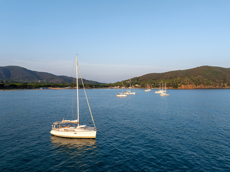 Aerial view of a sailboat floating in the calm blue waters of Lacona Bay surrounded by green hills and a clear sky in Lacona Tuscany Italy.