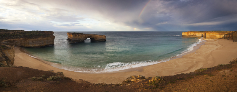 View of the London Arch and limestone cliffs along a sandy beach with a rainbow in the sky at Twelve Apostles Marine National Park Australia. View of the London Arch and limestone cliffs along a sandy beach with a rainbow in the sky at Twelve Apostles Marine National Park Australia.