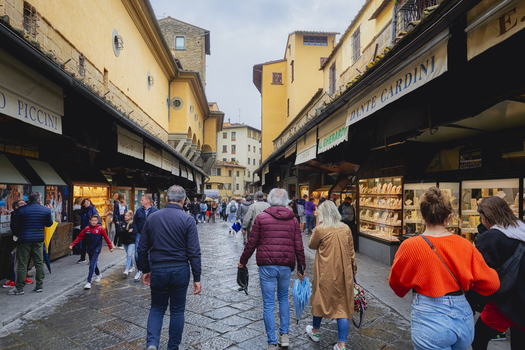 Florence Italy 05 May 2022 View of the historic Ponte Vecchio bridge featuring iconic jewelry shops stone paved walkway and crowds of people walking under yellow buildings. Florence Italy 05 May 2022 View of the historic Ponte Vecchio bridge featuring iconic jewelry shops stone paved walkway and crowds of people walking under yellow buildings.
