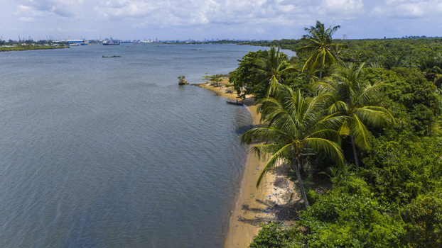 Aerial view of the lush palm trees and sandy shoreline along the Badagry Creek with a distant city skyline under a cloudy sky in Badagry Lagos Nigeria.