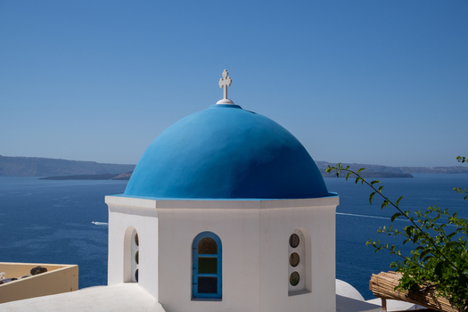 View of the Three Bells of Fira blue domed church overlooking the Aegean Sea and volcanic caldera under a clear blue sky in Fira Greece. View of the Three Bells of Fira blue domed church overlooking the Aegean Sea and volcanic caldera under a clear blue sky in Fira Greece.