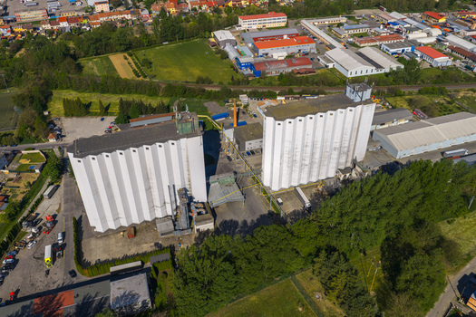 Aerial view of large white industrial grain silos and warehouse buildings surrounded by green trees and urban structures in Kyjov South Moravian Region Czechia.